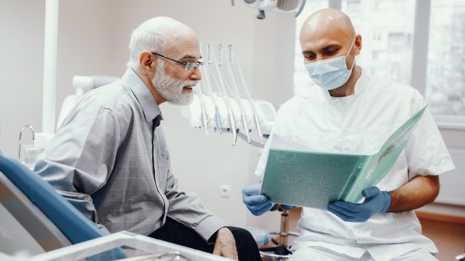 homem idoso sentado no consultório do dentista ao lado de um dentista com máscara e um documento